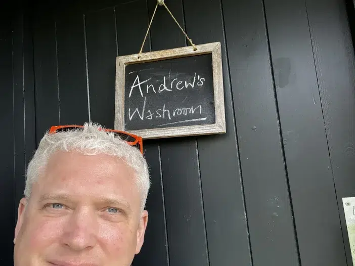 A man with short grey hair stands in front of a dark door with a chalkboard sign that reads "Andrew's Loo.